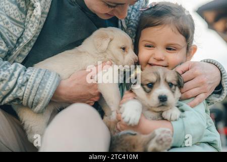 Porträt einer jungen Mutter mit ihrer kleinen Tochter, die mit Welpen spielt, draußen im Garten. Familie und Haustier zu Hause. Stockfoto