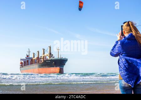 Frau filmt auf ihrem Handy ein großes Frachtschiff an der Küste, das auf Grund gelaufen ist, und einen Kitesurfer. Vityazevo, Russland - 12.02.2023 Stockfoto