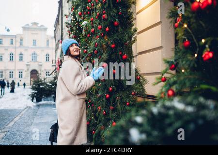 Weihnachtsstimmung. Glückliche Frau, die auf der Stadtstraße an Restaurants vorbeiläuft, die mit Tannengirlande und Ornamenten für Neujahr unter fallendem Schnee dekoriert sind Stockfoto