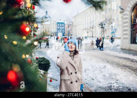 Porträt einer stilvollen Frau, die auf der zu Weihnachten dekorierten Stadtstraße unter fallendem Schnee läuft und Architektur genießt. Wintermode Stockfoto