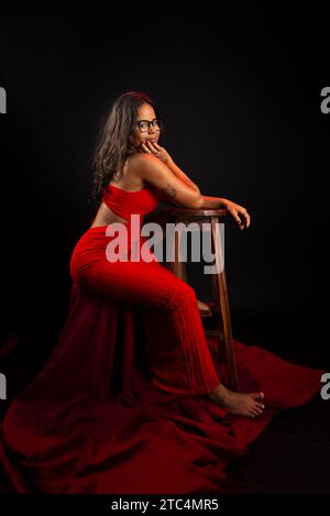 Schöne junge Frau in rot gekleidet, die mit der Hand auf dem Kinn sitzt und eine Brille trägt. Isoliert auf schwarzem Hintergrund. Stockfoto