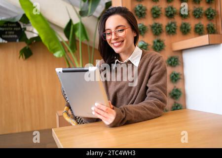 Lächelnde Frau, die ein digitales Tablet in der Hand hält. Selbstbewusste Frau sitzt im Flur und blickt auf die Kamera, die ein digitales Tablet vor unscharfem Hintergrund hält. Die Frau verwendet das Tablet für die Arbeit Stockfoto