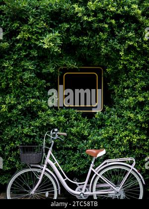 Quadratisches schwarzes, leeres Ladenschild, umgeben von grünem Busch mit Vintage-Fahrrad, vertikaler Stil. Gelbes Vorzeigetafel im Restaurant, Geschäft oder CA Stockfoto
