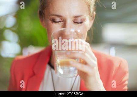 Nachhaltiger Arbeitsplatz. Lächelnde, moderne Eigentümerin eines kleinen Unternehmens bei der Arbeit in einer roten Jacke mit einer Tasse Wasser. Stockfoto