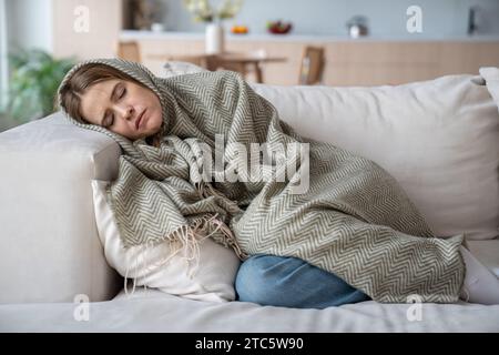 Kranke Frau, die auf dem Sofa liegt, mit geschlossenen Augen in kariertem Schüttelfrost, Erkältung, Grippe, Virus, Entzündung eingewickelt Stockfoto