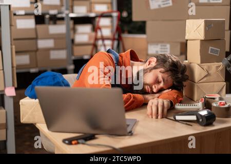 Erschöpfter Mann, der auf seinem Schreibtisch in der Nähe eines Laptops schläft, müde von Überarbeit im Lagerhaus. Workaholic leiden unter chronischer Müdigkeit Stockfoto