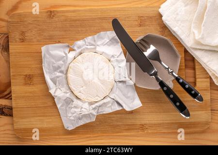 Duftender Brie-Käse mit Papierverpackung, Gabel und Messer auf hölzernem Küchenbrett, Makro, Draufsicht. Stockfoto