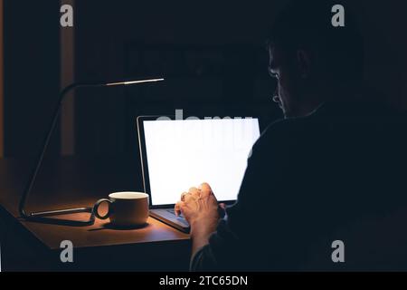 Ein Mann sitzt in einem dunklen Raum vor einem leeren Laptop-Bildschirm. Stockfoto