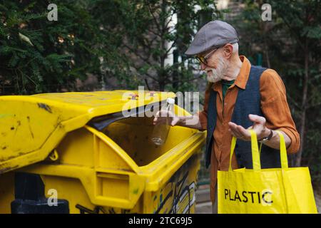 Senior Mann wirft Plastikmüll, Flaschen in den Recyclingbehälter vor der Wohnung. Ein älterer Mann sortiert den Abfall nach Material Stockfoto