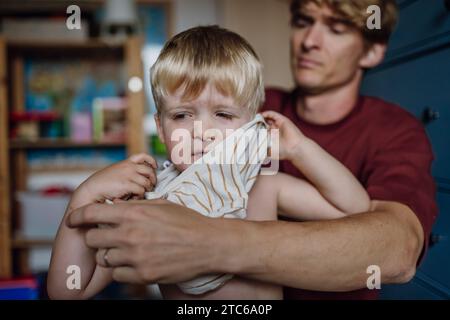 Vater zieht Hemd auf den kleinen Sohn und zieht sich morgens vom Schlafanzug. Der Junge ist traurig, weinend. Stockfoto