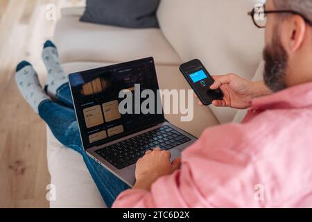 Mann mit Notebook und Smartphone auf Sofa, Online-Banking, Internetbanking zu Hause über Website und App. Online-Bezahlung mit Smartphone Stockfoto