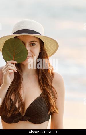 Entspannte Brünette in schwarzem Bikini und Sonnenhut, Blatt halten, bei Sonnenuntergang. Stockfoto