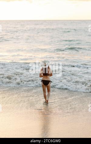 Besinnliche junge Frau in schwarzem Bikini geht bei Sonnenuntergang am Strand. Stockfoto