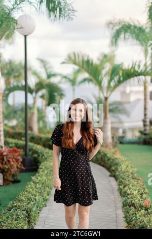 Lächelnde junge Frau in schwarzem Kleid schlendert im Dominikanischen Strandresort Stockfoto
