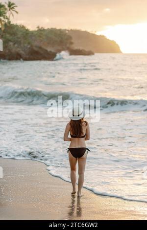 Junge Frau in Sonnenhut und Bikini spaziert am Dominikanischen Strand bei Sonnenuntergang. Stockfoto