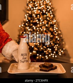 Die Weihnachtsmann-Hand greift nach der Milchflasche mit Brownies auf dem Teller Stockfoto