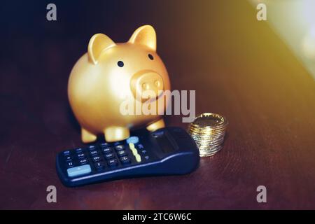 Eine goldene Schweinebank auf einem schwarzen Rechner mit Münzen auf einem dunklen Holztisch. Sparkonzepte für Finanzen, Geld und Wirtschaft. Stockfoto
