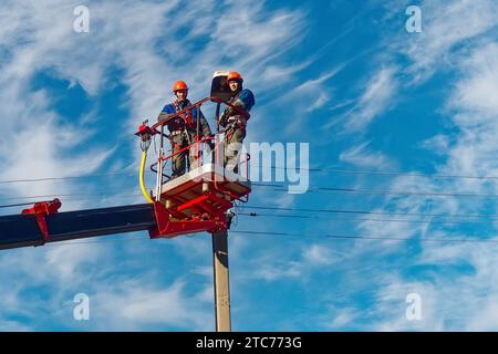 Zwei Elektriker von der Halterung der Antennenplattform oder des Krans reparieren die Straßenbeleuchtung. Professionelle Elektriker tragen Helme, Overalls und Versicherungsarbeiten in der Höhe. Ansicht der Arbeiter von unten nach oben zum Himmel. Authentischer Workflow. Stockfoto