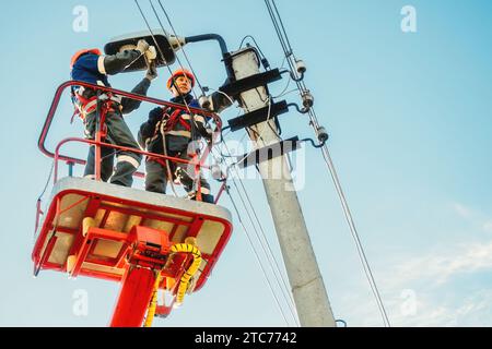 Zwei Elektriker von der Halterung der Antennenplattform oder des Krans reparieren die Straßenbeleuchtung. Professionelle Elektriker tragen Helme, Overalls und Versicherungsarbeiten in der Höhe. Ansicht der Arbeiter von unten nach oben zum Himmel. Authentischer Workflow. Stockfoto