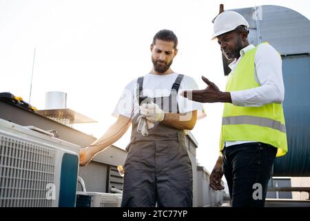 Kaukasische bärtige Konstruktion stehend und mit Messgerät für Klimaanlage, während Afro-Kollege im Freien positive Ergebnisse bestätigt. Conce Stockfoto