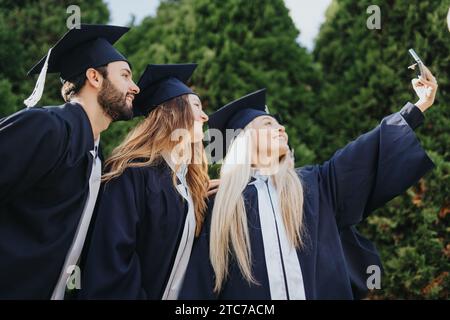 Multiethnische Hochschulabschlüsse feiern sonnigen Tag im Park. Sie lächeln, reden und werfen Mützen, nehmen Selfies und schätzen ihre positiven Erinnerungen Stockfoto