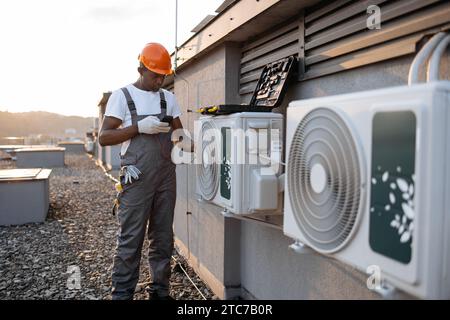 Ein vielbeschäftigter Bauingenieur in grauem Anzug hält ein Digitalmultimeter und überprüft den Spannungspegel bei der Handhabung von Klimaanlagen im Freien. Afrikanisch-amerikanisch Stockfoto