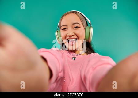 Fröhliche und junge asiatische Frau in schnurlosen Kopfhörern, Musik hören und Selfie auf Türkis machen Stockfoto