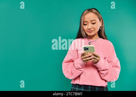 Porträt einer fröhlichen asiatischen Frau in rosa Sweatshirt mit Smartphone vor türkisfarbenem Hintergrund Stockfoto