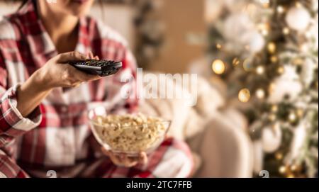 Eine Frau im Pyjama hält während der Weihnachtsfeiertage eine Fernbedienung in der einen Hand, eine volle Schüssel Popcorn in der anderen Hand – NAHAUFNAHME. Stockfoto