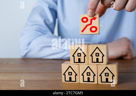 Steigende Hypothekenzinsen, dargestellt durch Prozentzeichen mit Aufwärtspfeil. Mann baut Pyramide aus Würfeln mit Häusersymbolen an Holztisch, Nahaufnahme Stockfoto