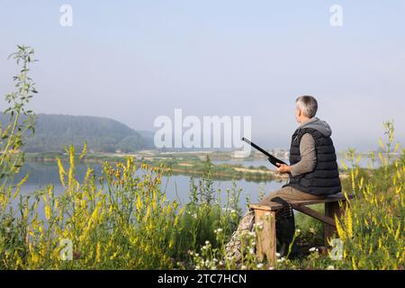 Mann mit Jagdgewehr sitzt auf einer Holzbank in der Nähe des Sees. Leerzeichen für Text Stockfoto