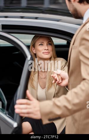 Verschwommener Mann, der die Hand einer lächelnden blonden Geschäftsfrau in formeller Kleidung hält, die aus einem Luxusauto rauskommt Stockfoto