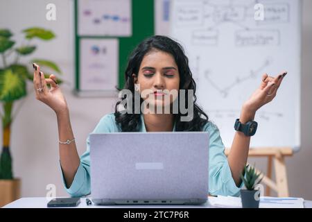 Frustrierte Geschäftsfrau oder Angestellte im Büro, die Meditation im Büro macht, während sie am Laptop arbeitet – Konzept von Achtsamkeit, Selbstpflege oder Wellness und Stockfoto