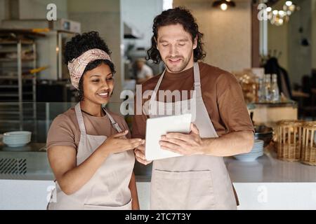 Zwei birassische Bäckerei-Mitarbeiter diskutieren Informationen auf einem digitalen Tablet-Bildschirm Stockfoto