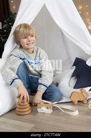 Ein hübscher lächelnder Junge sitzt in einem Wigwam-Zelt mit Holzspielzeug in der Nähe. Er sieht träumerisch und fröhlich zur Seite. Kinderzimmer, Kindheit Stockfoto