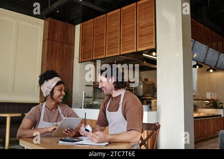 Birassische Kollegen, die sich fröhlich anschauen und Gewinn zählen, die am Tisch in der Bäckerei sitzen Stockfoto