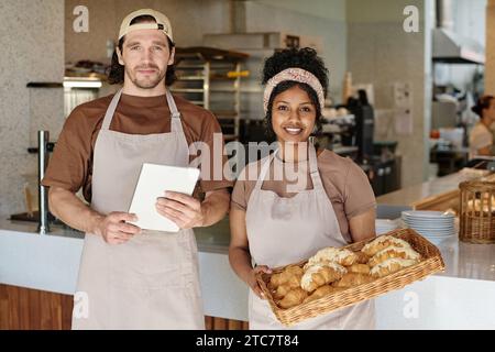 Mittelgroße Aufnahme junger birassischer Bäckereiarbeiter, die gegen die Theke stehen und in die Kamera lächeln Stockfoto