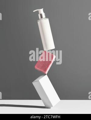 Creative Concept Körperpflege. Weiße Flasche, Stück Seife und Box für Kosmetikprodukte, die vor grauem Hintergrund aufeinander balanciert sind. Stockfoto