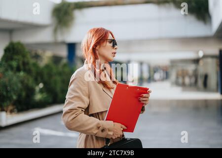Rothaarige Geschäftsfrau, die in der Stadt arbeitet. Sie hält eine Zwischenablage mit Papierkram drauf. Stockfoto
