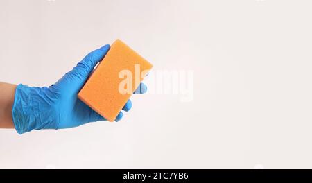 Orangefarbener Geschirrspülschwamm in der Hand einer Frau auf grauem Hintergrund. Eine Hand in einem latexblauen Handschuh hält einen Schwamm für die Nassreinigung. Professionelle Reinigung. Stockfoto