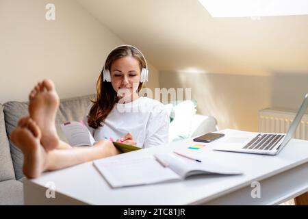 Selbstentwicklung. Attraktive brünette Frau in Kopfhörern, die ein Arbeitsbuch am Schreibtisch verwendet. Selbstgesteuertes Lernen. Stockfoto