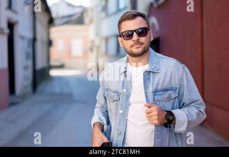 Junger erwachsener Mann mit Sonnenbrille und Jeansjacke posiert während er im Frühling auf der Straße steht. Stockfoto