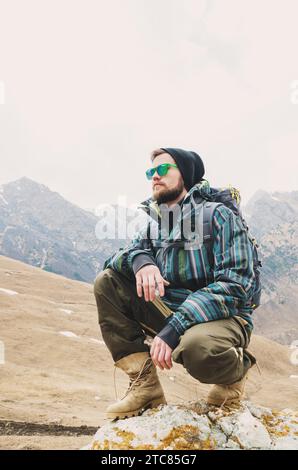 Müder bärtiger Hipster mit Fernglas in den Händen sitzt auf einem Stein zwischen den kaukasischen Bergen und blickt in die Ferne Stockfoto