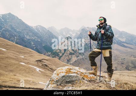 Müder bärtiger Hipster mit Fernglas in den Händen sitzt auf einem Stein zwischen den kaukasischen Bergen und blickt in die Ferne Stockfoto