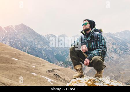 Müder bärtiger Hipster mit Fernglas in den Händen sitzt auf einem Stein zwischen den kaukasischen Bergen und blickt in die Ferne Stockfoto