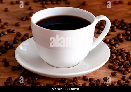 Eine weiße Porzellantasse mit schwarzem Kaffee steht auf einem Holztisch. Kaffeebohnen Stockfoto