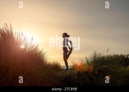 Sportliche Frau dehnt ihre Beine aus, macht stehende Quadrizeps-Oberschenkel-Dehnung draußen bei Sonnenaufgang Stockfoto