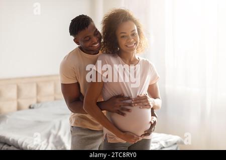 Afrikanischer Ehemann umarmt den Bauch seiner schwangeren Frau im Schlafzimmer Stockfoto