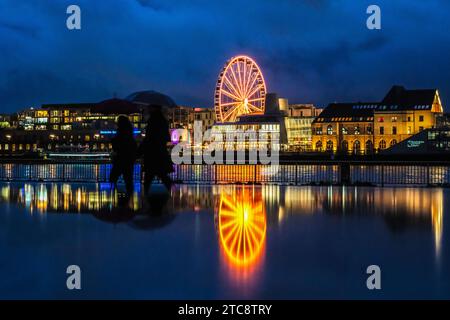Köln, Deutschland. Dezember 2023. Wanderer mit Regenschirmen passieren am Abend ein Riesenrad, das sich auf einem regendurchtränkten Weg am Rheinufer spiegelt. Quelle: Oliver Berg/dpa/Alamy Live News Stockfoto