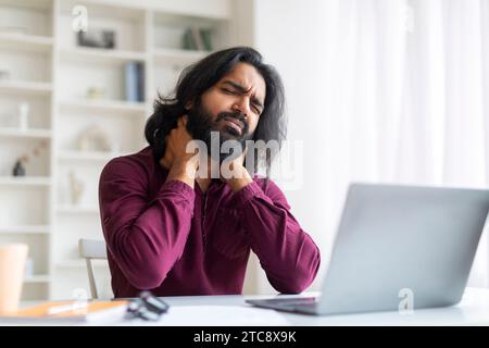 Indischer freiberuflicher Mann, der Nackenschmerzen empfindet, während er zu Hause mit einem Laptop arbeitet Stockfoto